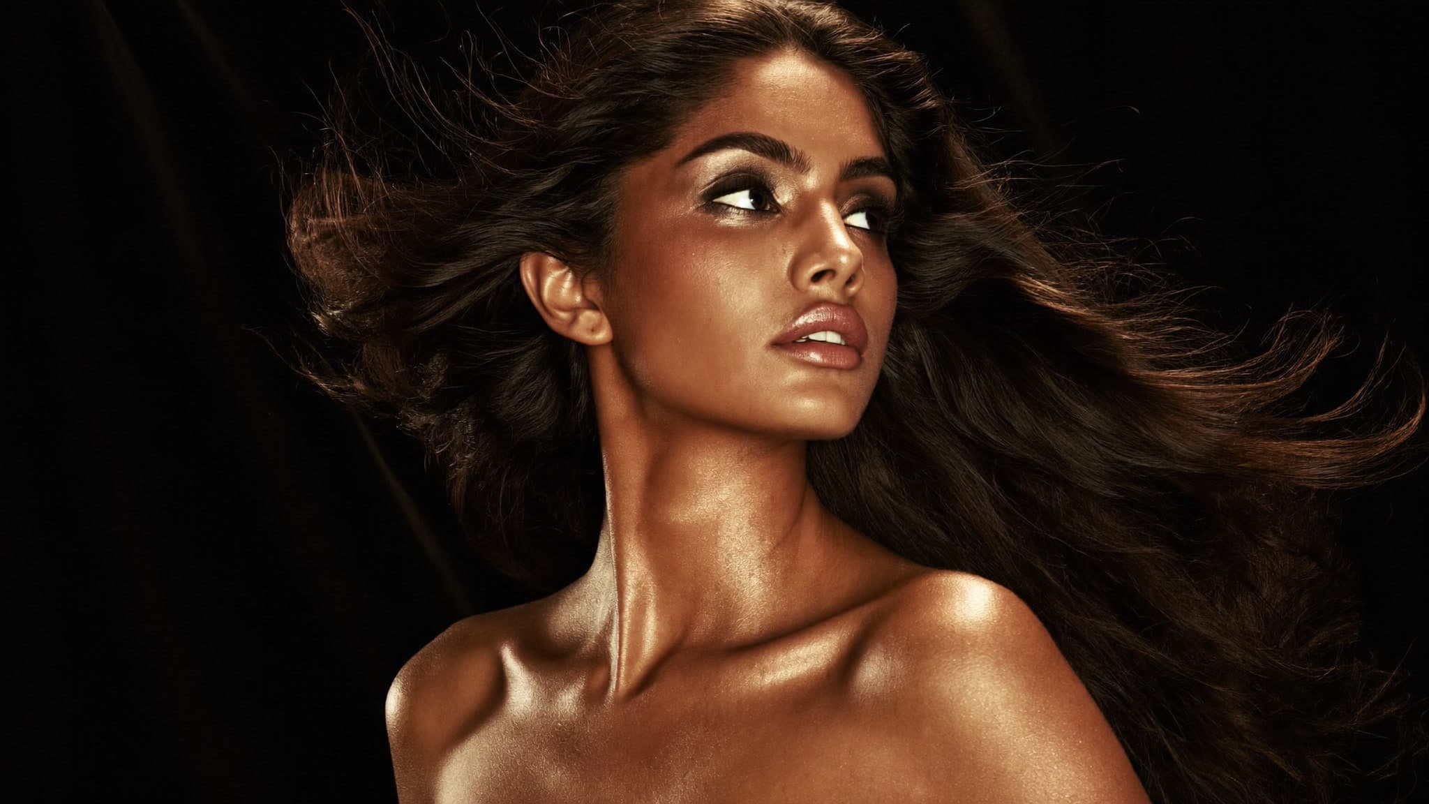 A model with windswept hair and radiant golden skin gazing upward in champagne-gold light — beauty campaign concept imagery